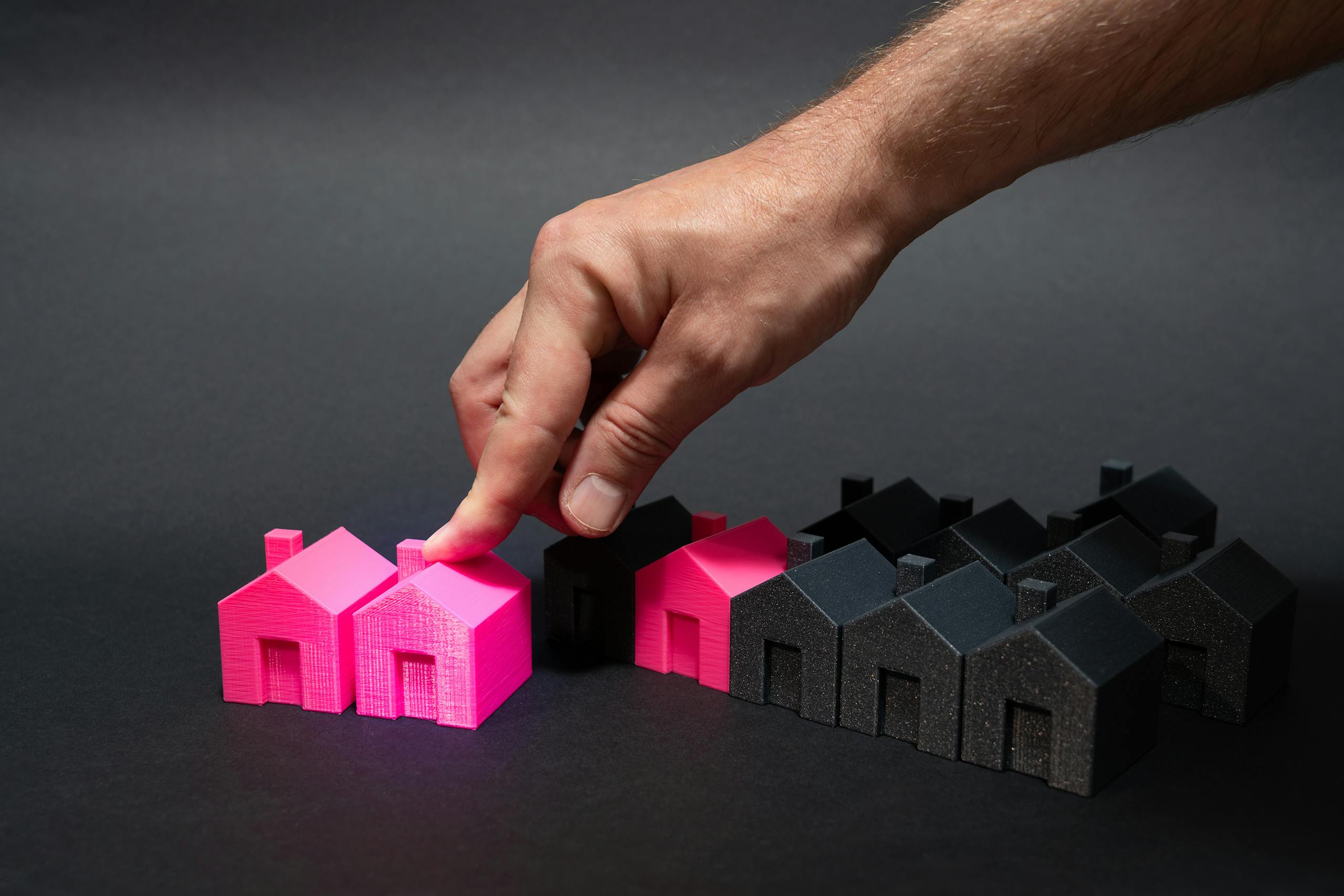 A hand choosing a vibrant pink house model among black miniatures, symbolizing real estate selection.