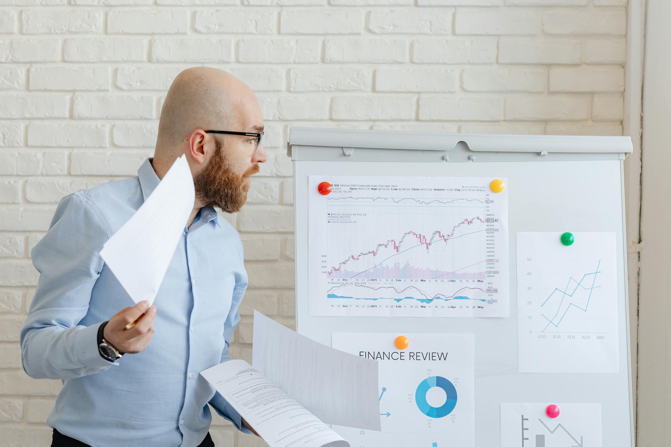 Bald businessman in smart casual attire analyzing financial charts on a whiteboard in an office setting.